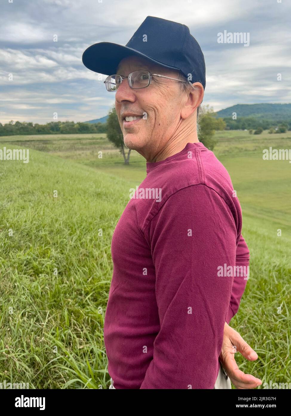 Active senior man hiker smiles at the top of a tall green grass hill. Stock Photo
