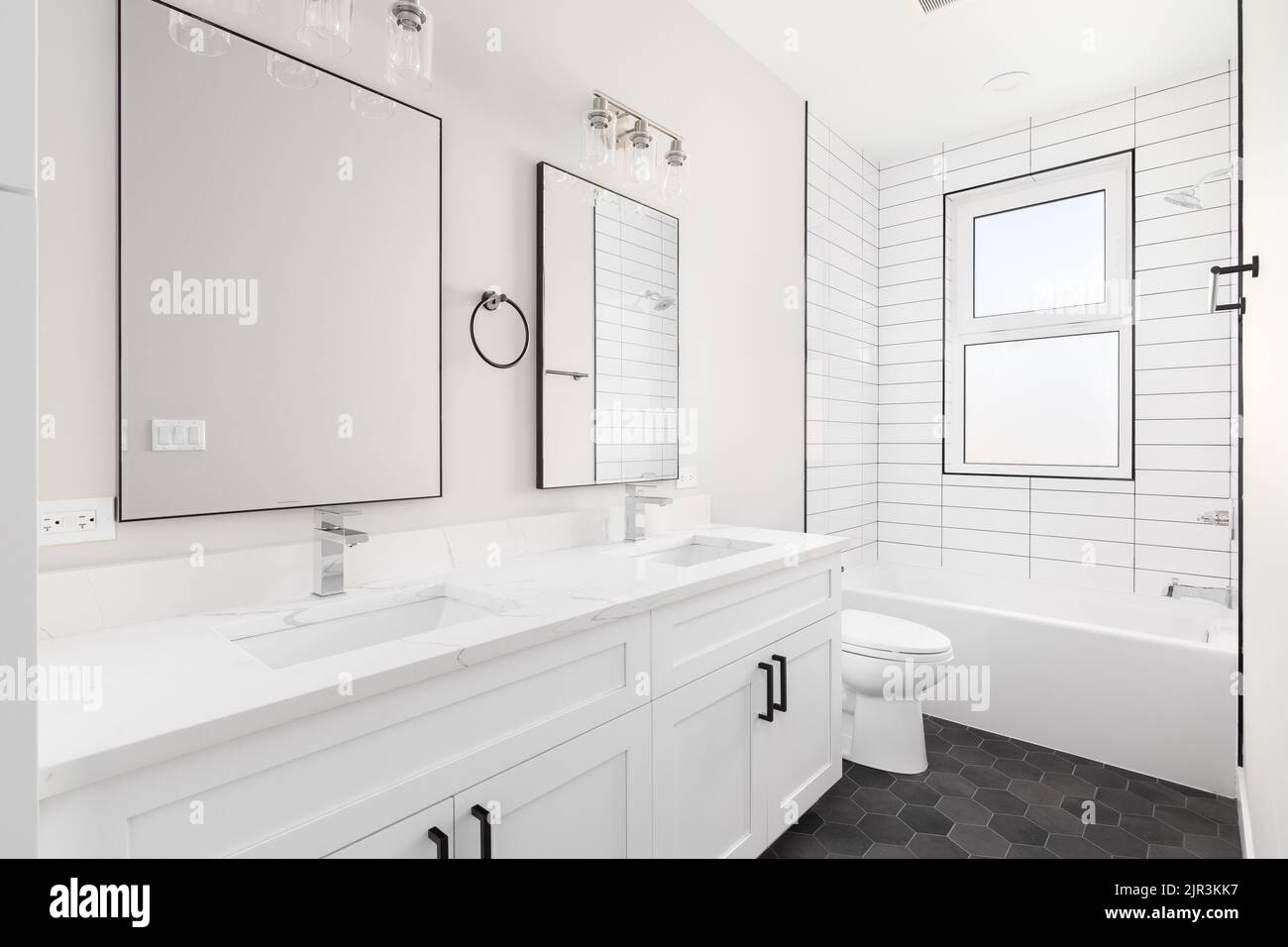 A white luxury bathroom with a white vanity cabinet and marble countertop, subway tile shower, and dark hexagon tiled flooring. Stock Photo
