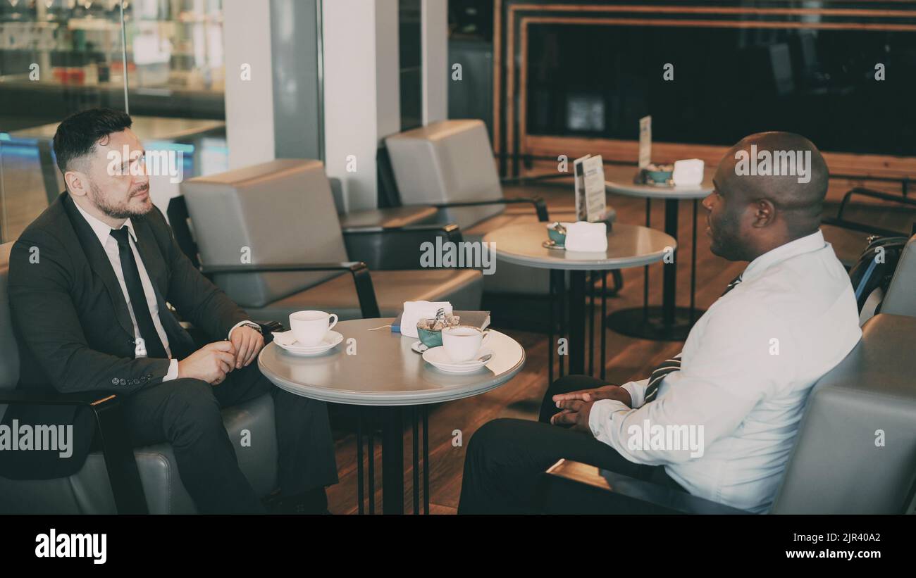 Two multi-ethnic businessmen sitting, smiling and discussing their startup in airy cafe during lunch time. Coffee cups are on their table Stock Photo