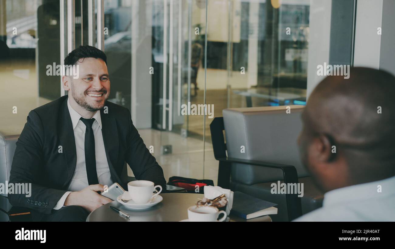 Two multi-ethnic businessmen sitting, smiling and discussing their startup in airy cafe during lunch time. Cheery African American partner strokes his knees happily. Coffee cups are on their table Stock Photo