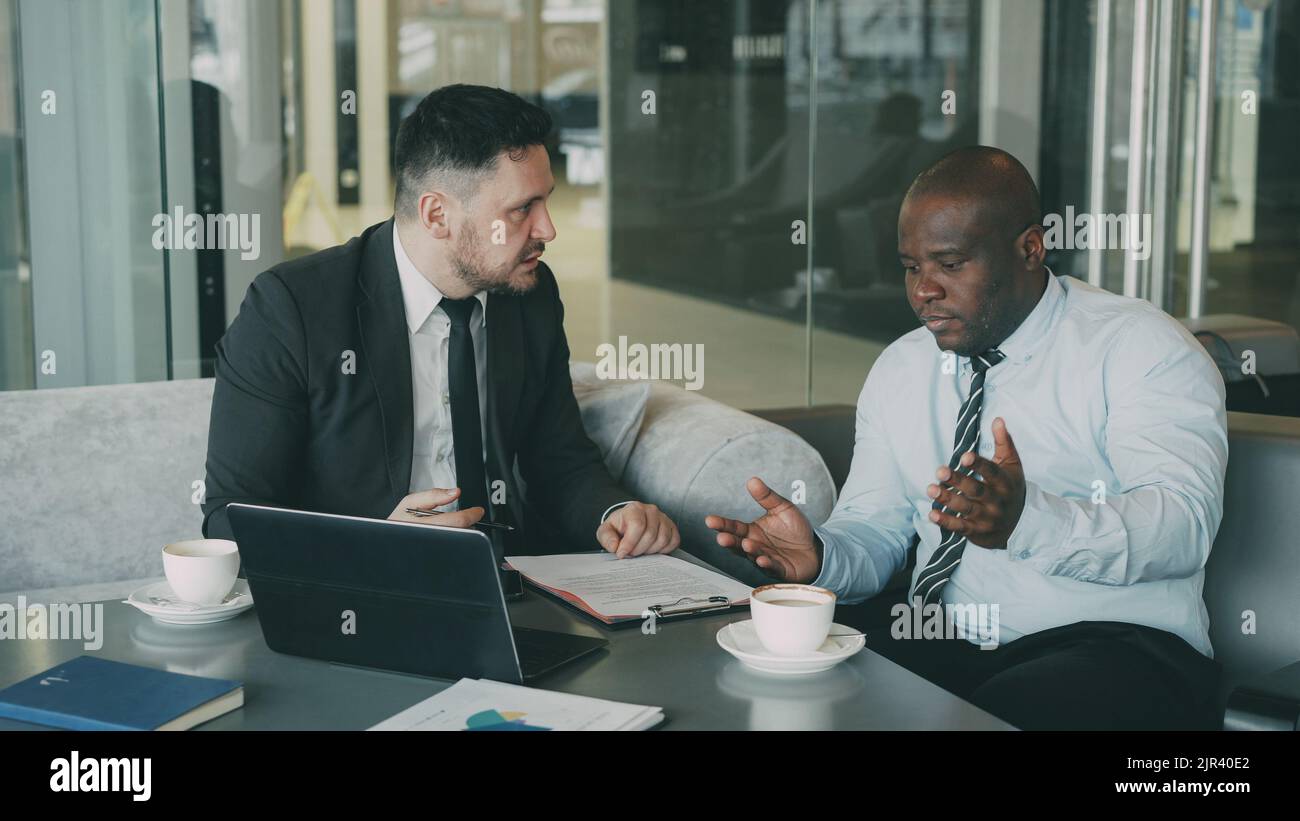 Angry Caucasian ceo in black business suit, white criticizes severely his African American employees during meeting in modern cafe. Irritated boss gesticulates emotionally and agressively Stock Photo