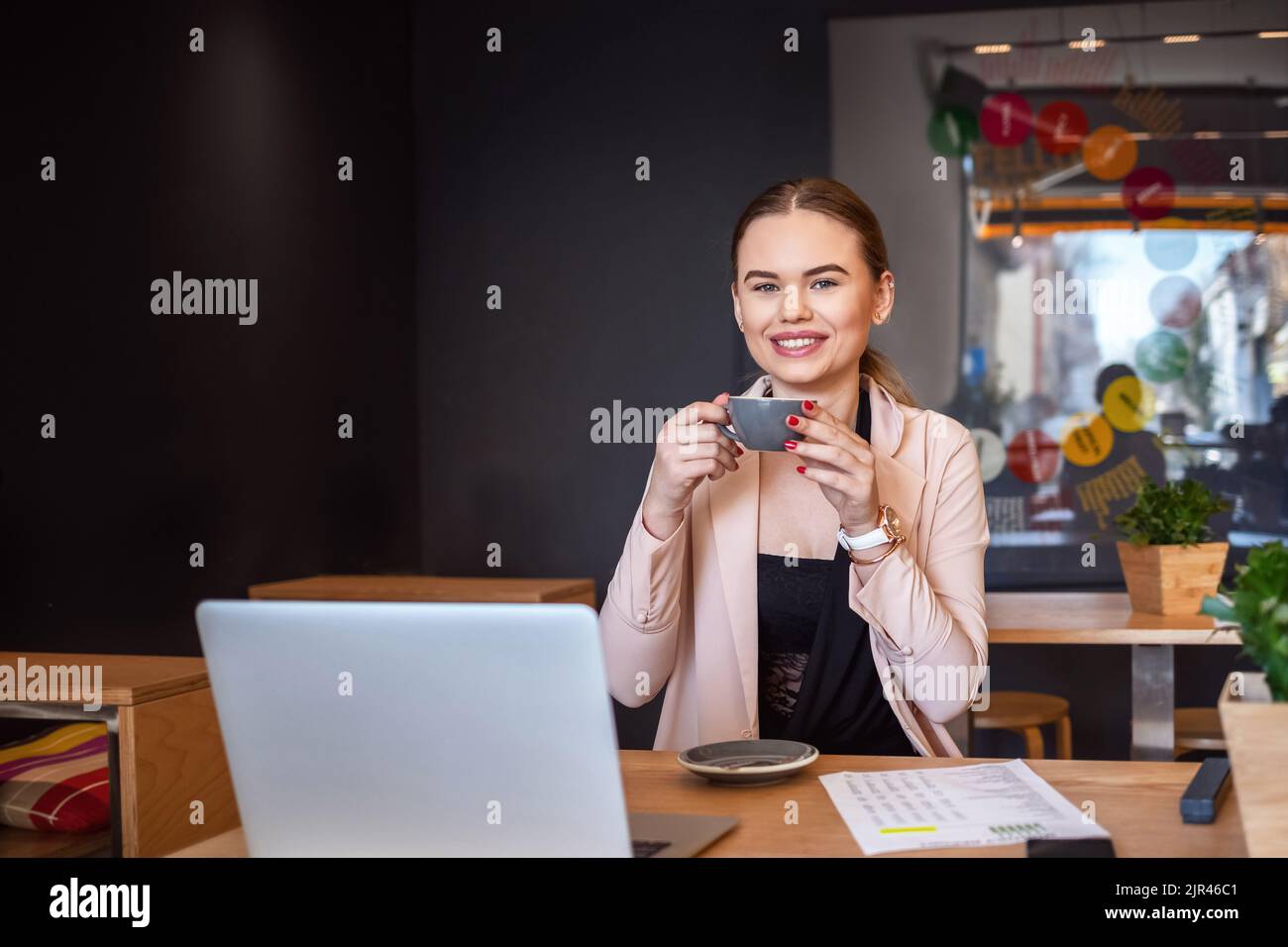 Smiling woman sitting in cafeteria holding coffee mug and working on laptop Stock Photo