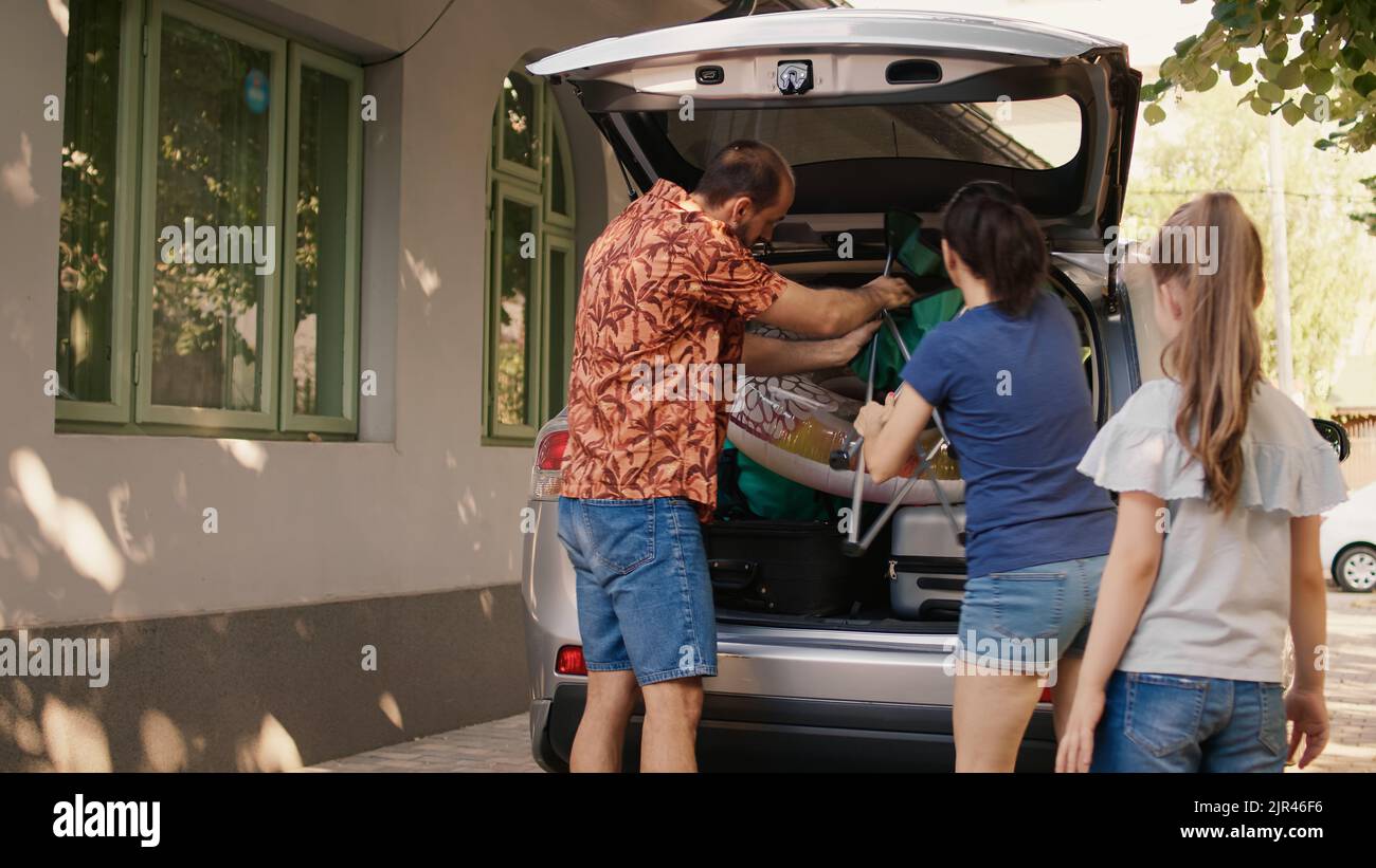 Family loading car trunk with field trip baggage while getting ready for summer holiday citybreak. Caucasian people putting voyage luggage and trolleys in vehicle while going on weekend vacation. Stock Photo