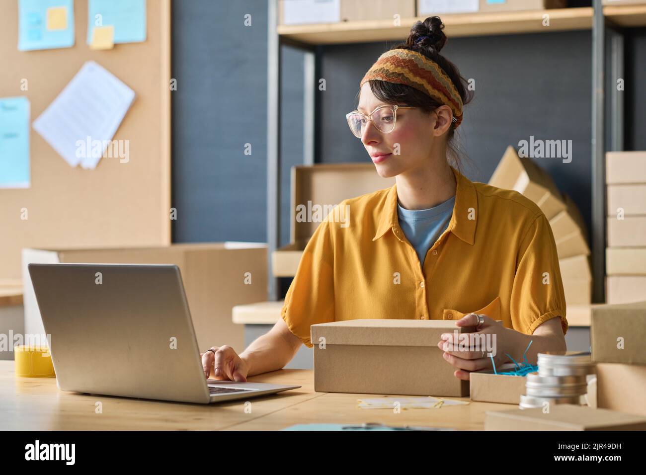 Warehouse woman sitting at table with laptop and cardboard box, and working with online orders to make parcels Stock Photo