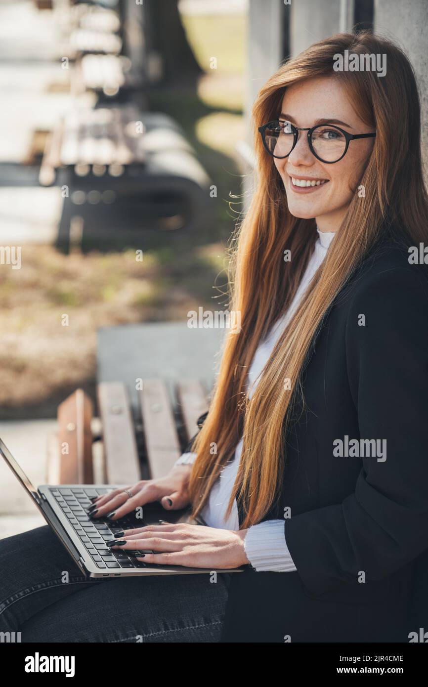 Read-headed student woman in black jacket sitting on bench in park outdoors, looking and smiling at camera use laptop. People urban lifestyle concept Stock Photo