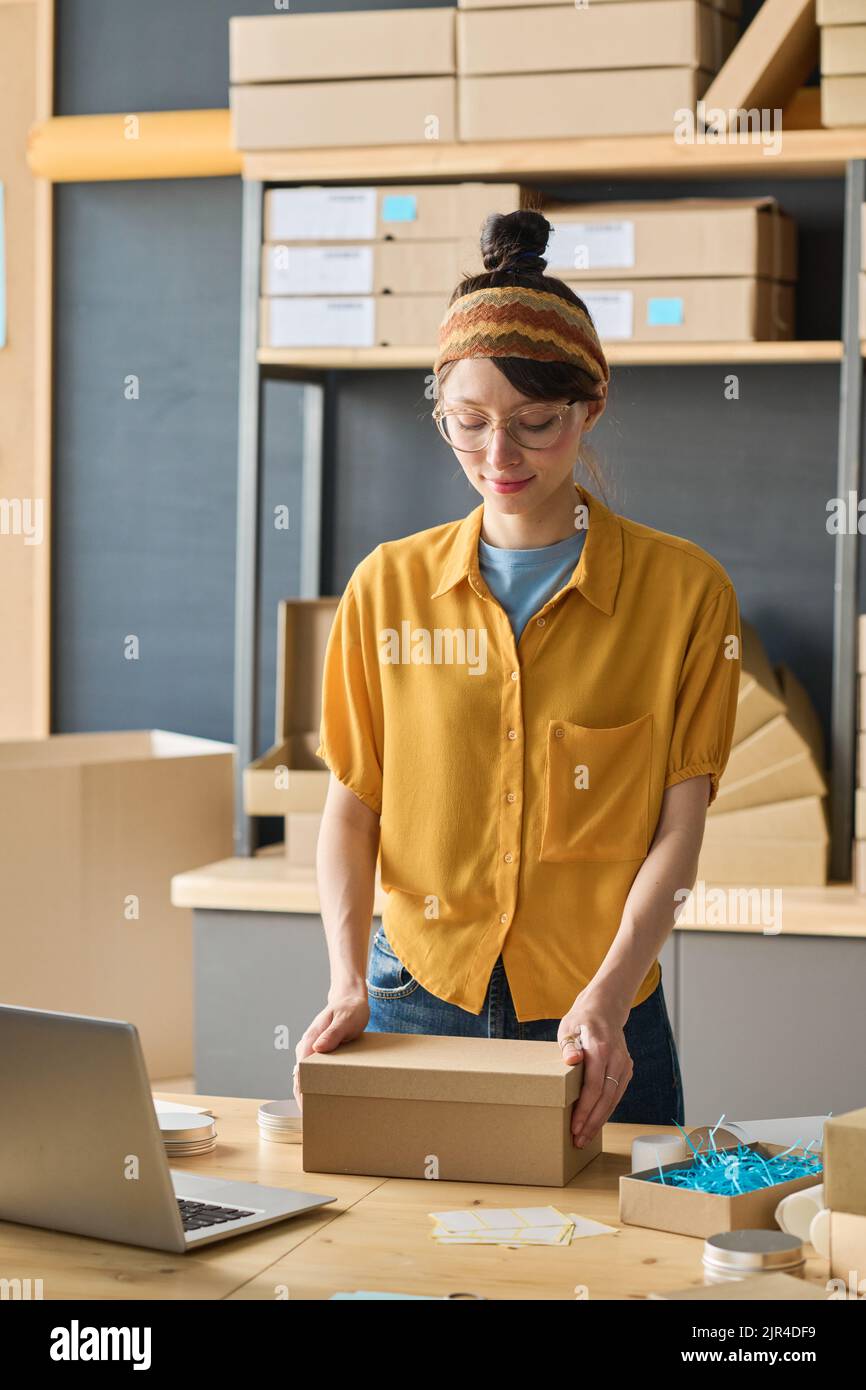 Young female worker packing goods in cadboard boxes at table in warehouse before delivery Stock Photo