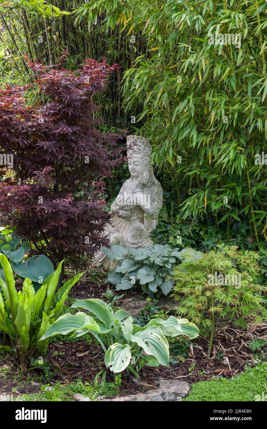 A small zen garden with shrubs and plants surrounding a carved stone figure; Gayton, Northamptonshire, UK Stock Photo