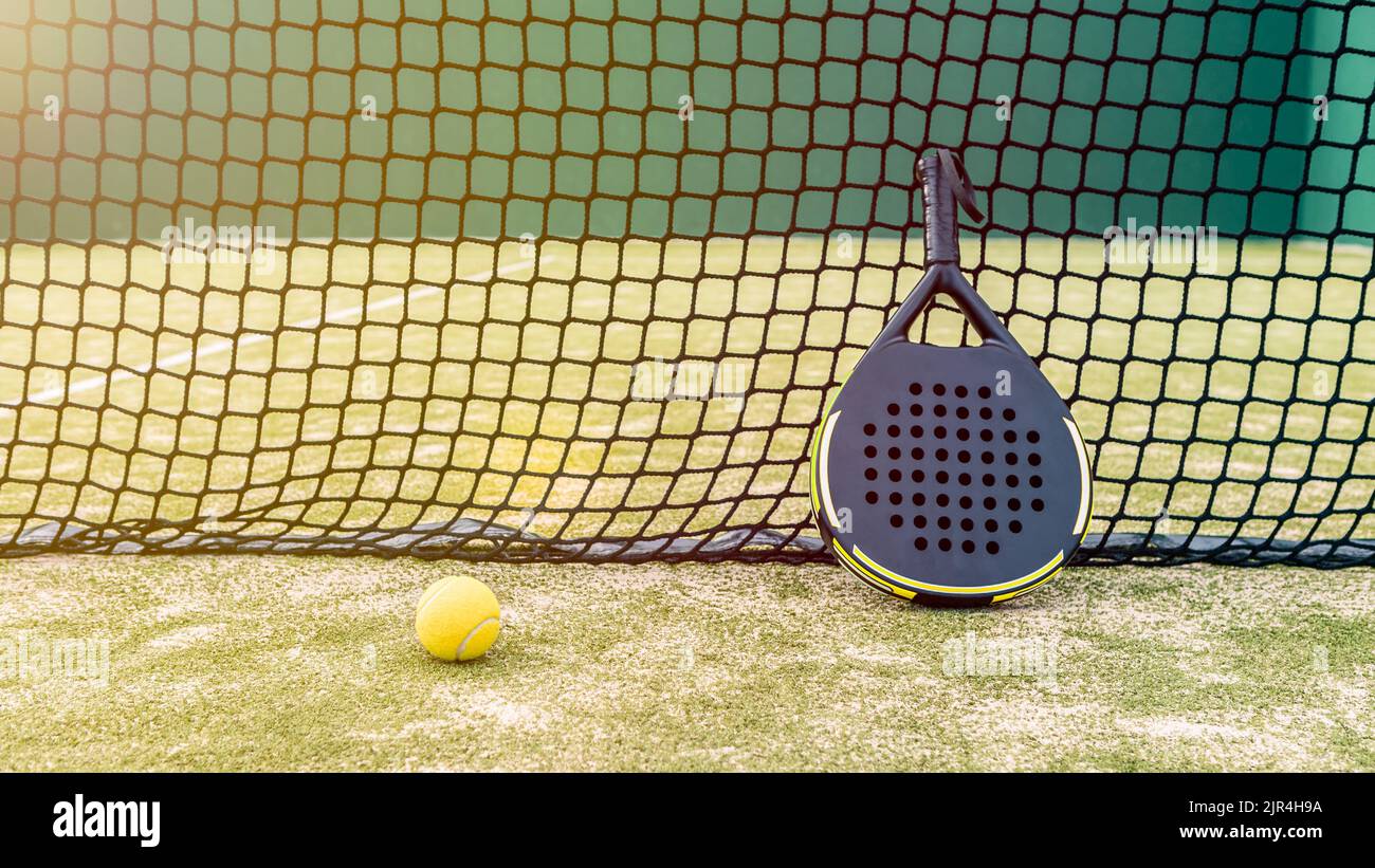 Padel racket and yellow ball behind net on a green court grass turf outdoors at sunset. Paddle is a racquet game. Professional sport concept with spac Stock Photo