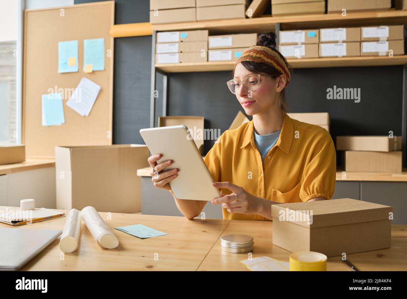 Young female worker tracking numbers of parcels on digital tablet while sitting at table with boxes in warehouse Stock Photo