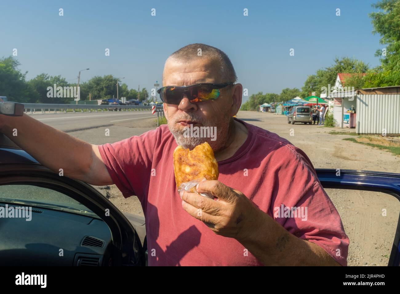Portrait of hungry Ukrainian senior driver taking patty near his car while standing near Vasylivka, Zaporizhzhia Oblast, Ukraine Stock Photo