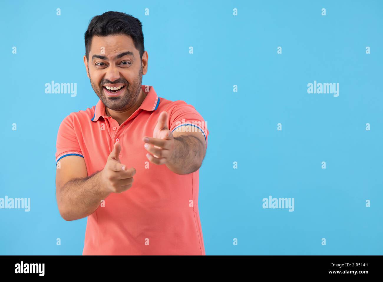 Smiling young man looking and pointing at camera against blue background Stock Photo