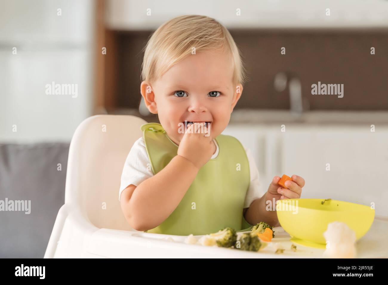 Happy blond baby in green bib eating with pleasure organic steamed