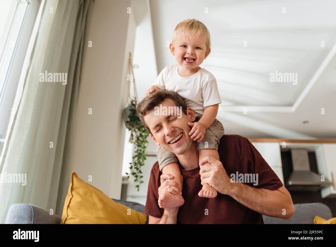 Laughing father giving his kid piggyback ride on shoulders, baby boy pulling daddy's hair, father and son spending time together on sunday morning in living-room. Dad playing with his toddler boy Stock Photo