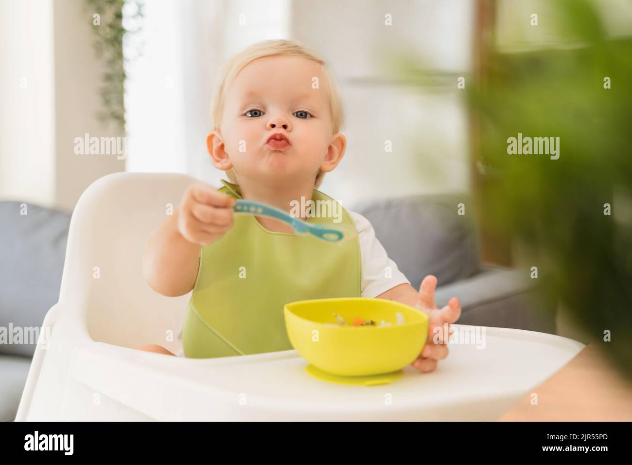 Adorable blond baby boy sitting at table in high chair with spoon in hand and yellow plate with breakfast in front of him, training skills to feed himself, wearing green bib. Baby eating by himself Stock Photo