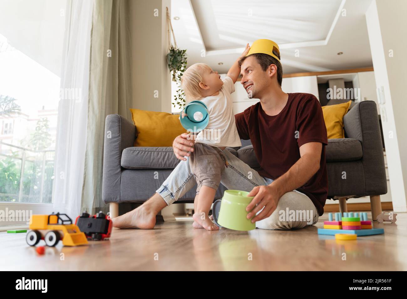 Young father and son having fun together. Blond baby toddler putting toy on dad's head sitting on floor in living-room, daddy entertaining his little boy, playing toys, active games at home Stock Photo