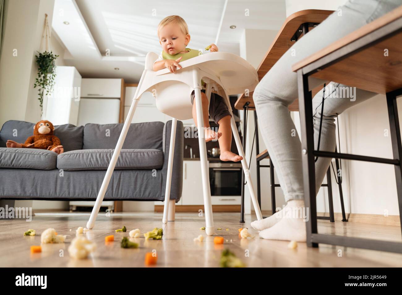 Side view of cute caucasian baby sitting in high chair with piece of vegetable looking with interest on messy and dirty floor covered with broccoli, carrot and cauliflower. Baby feeding concept Stock Photo