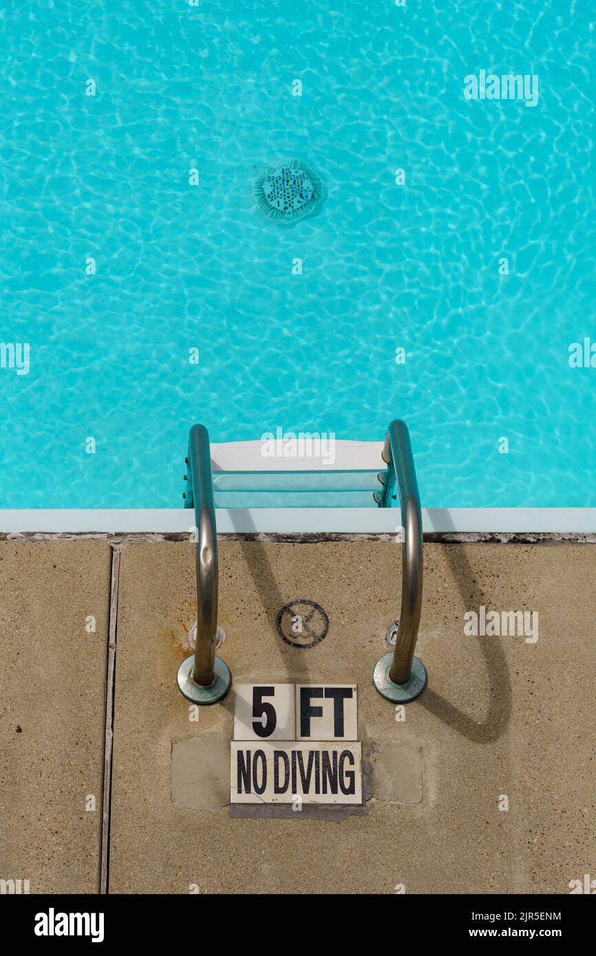 No diving sign by a pool ladder. The water is clear and blue, and you can see the pool drain. Water depth is marked as 5 feet. Stock Photo