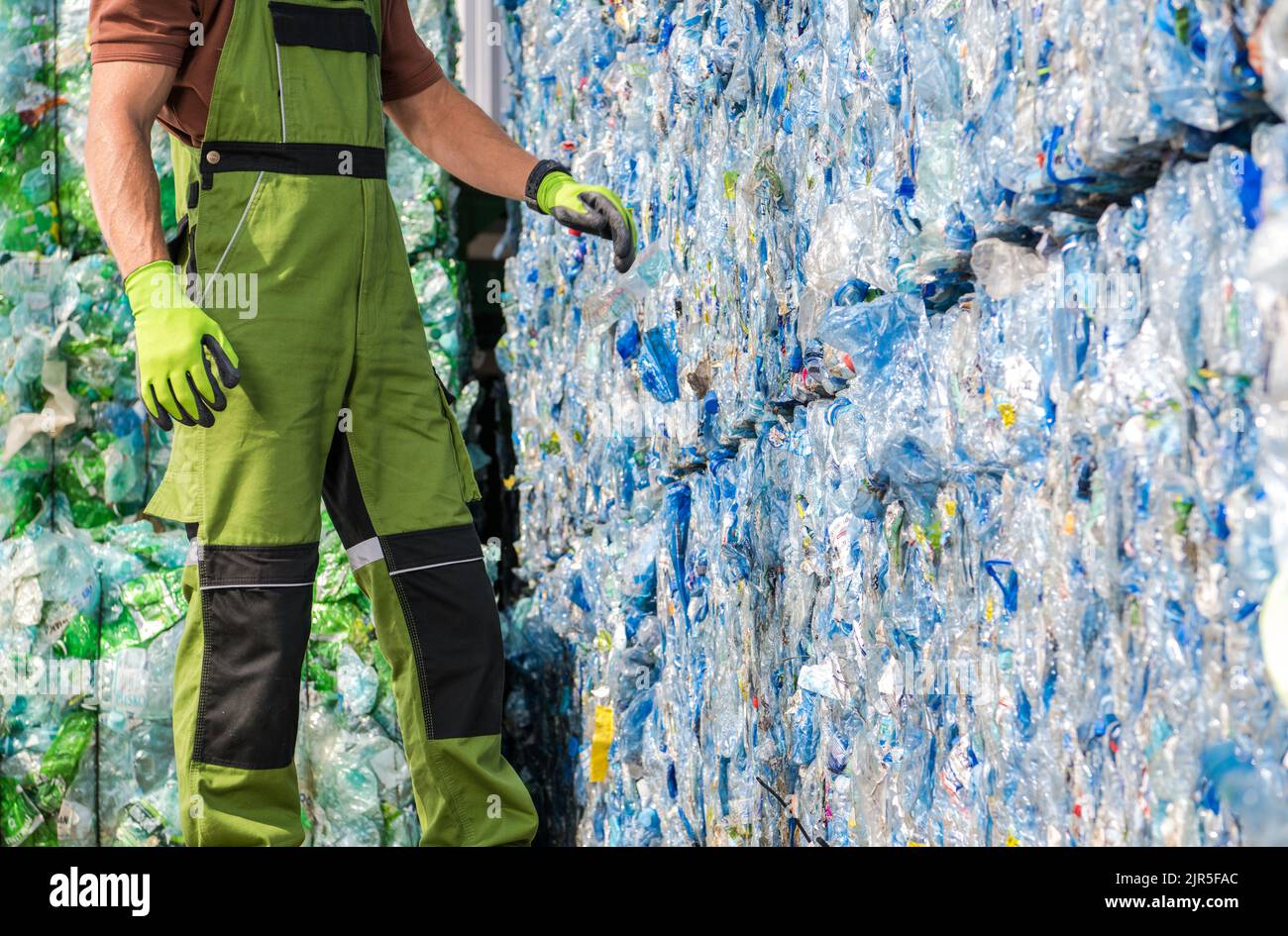 Caucasian Waste Management Worker in Front of a Pile of Pressed Plastic PET Bottles. Trash Sorting Facility. Stock Photo
