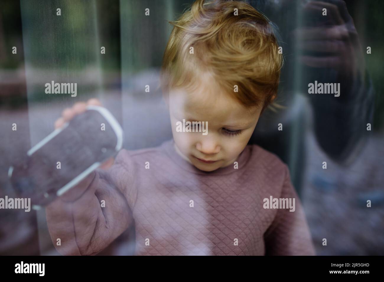 Little child standing alone with car toy behind the window, photo trough glass. Stock Photo