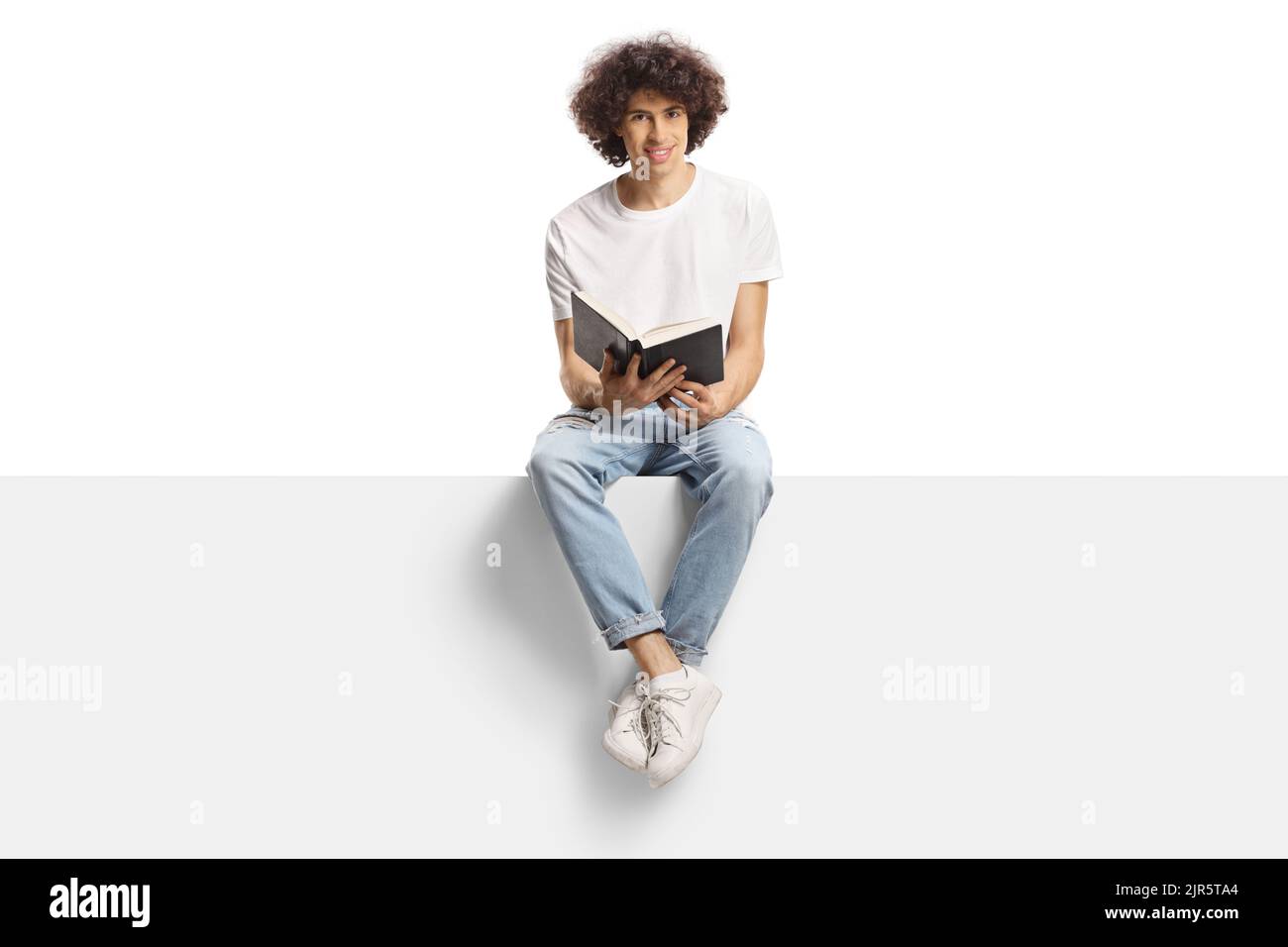 Young man with curly hair sitting on a panel and holding an open book isolated on white background Stock Photo