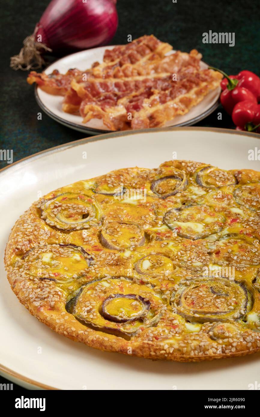 Homemade onion omelette in a white dish, crispy bacon slices and red chili peppers on the background. Stock Photo