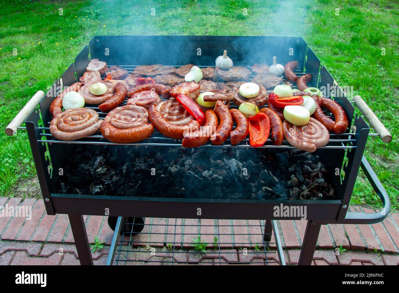 Assorted types of grilled meat with vegetables on the barbecue mobile grill with sausage, weisswurst, red pepper, bacon, onion and garlic with smoke a Stock Photo