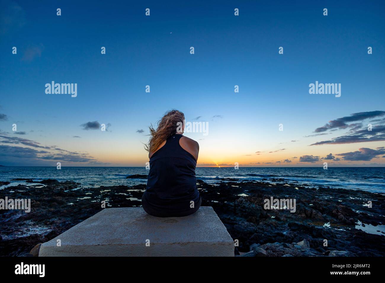 A back view of a female sitting on a shore and looking at calm sea ...