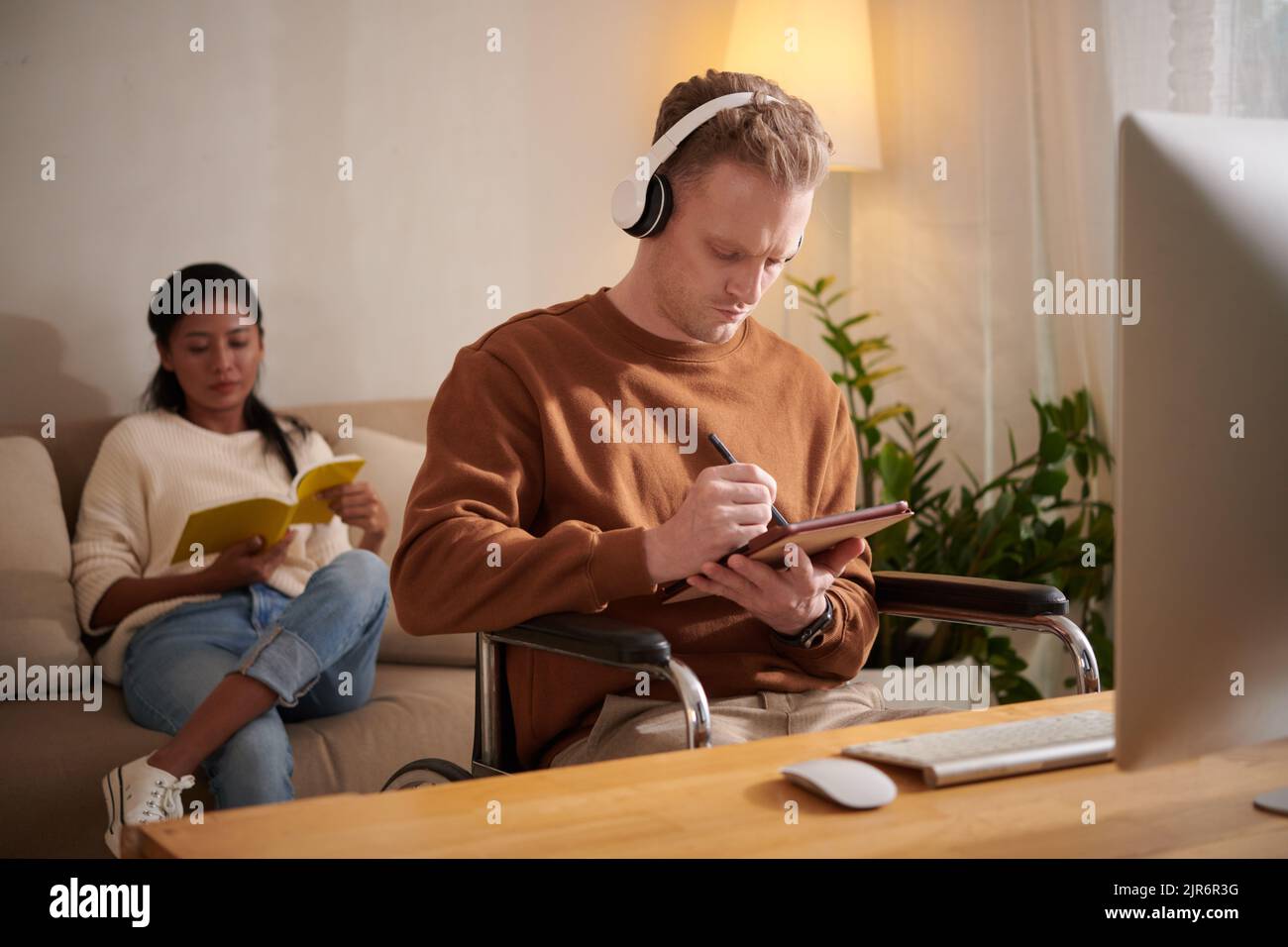 Young man with disability listening to music and working on tablet computer at home, girlfriend reading book in background Stock Photo