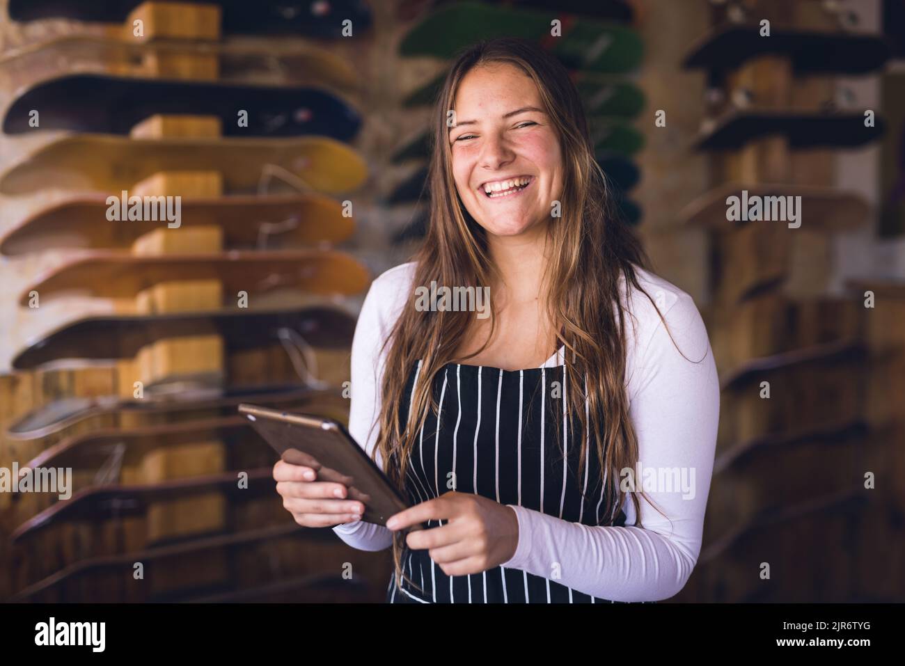 Image of happy caucasian woman in apron with tablet posing in skate shop Stock Photo