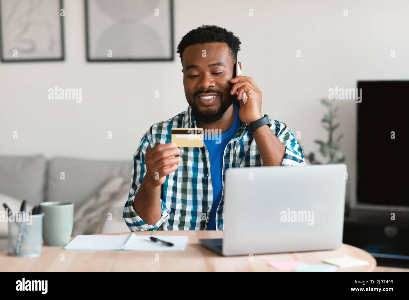 Cheerful Black Guy Holding Credit Card Talking On Phone Indoors Stock Photo
