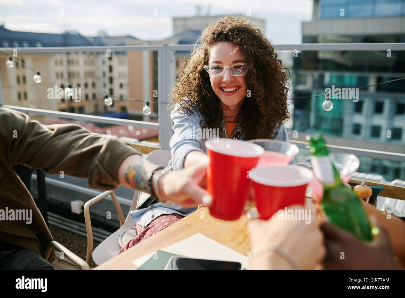 Young smiling woman with dark long curly hair toasting with drinks of her friends witting by table in rooftop cafe and having nice time Stock Photo