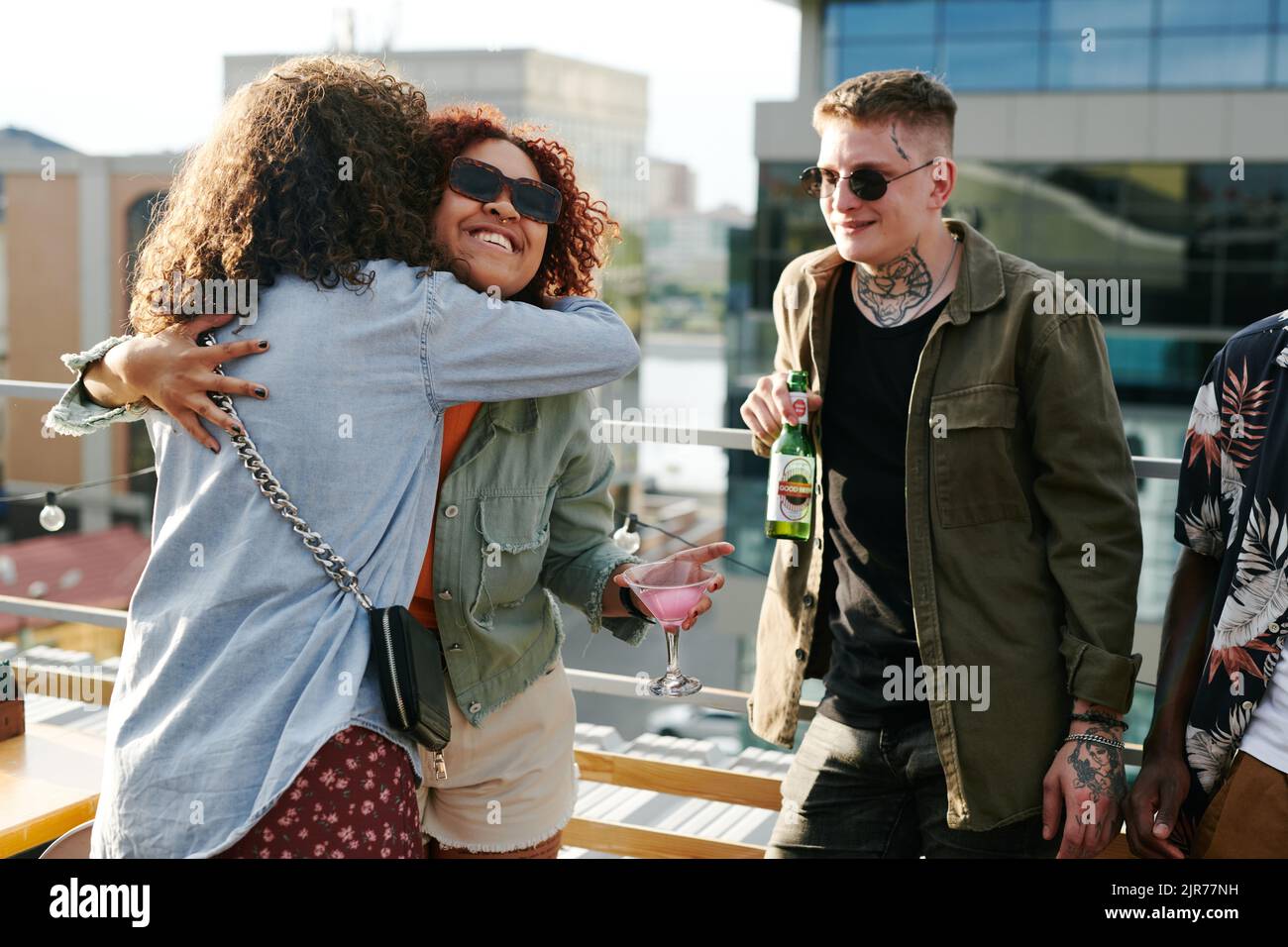 Happy young black woman with cocktail in martini glass giving hug to friend while standing against guy with bottle of beer Stock Photo