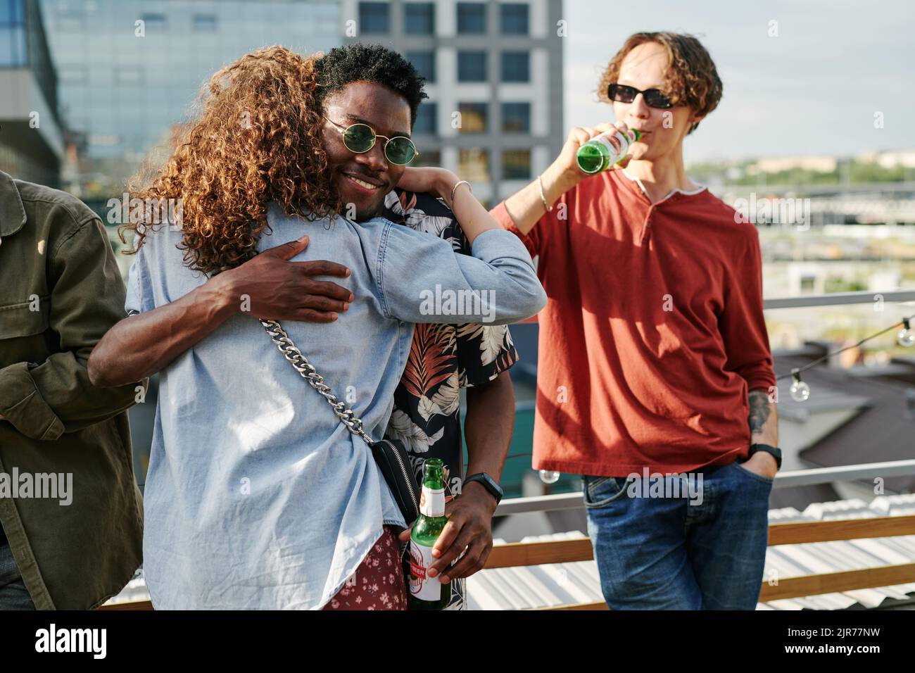 Happy young black man with bottle of beer embracing adolescent girl in denim jacket while greeting each other at rooftop party Stock Photo
