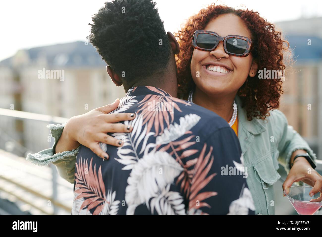 Young cheerful black woman in sunglasses giving hug to African American man while both enjoying rooftop party Stock Photo