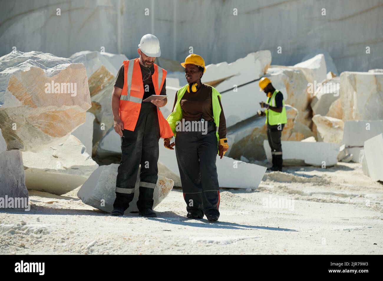 Two young intercultural workers of marble quarry discussing online manual guide while standing against heaps of huge rocks Stock Photo