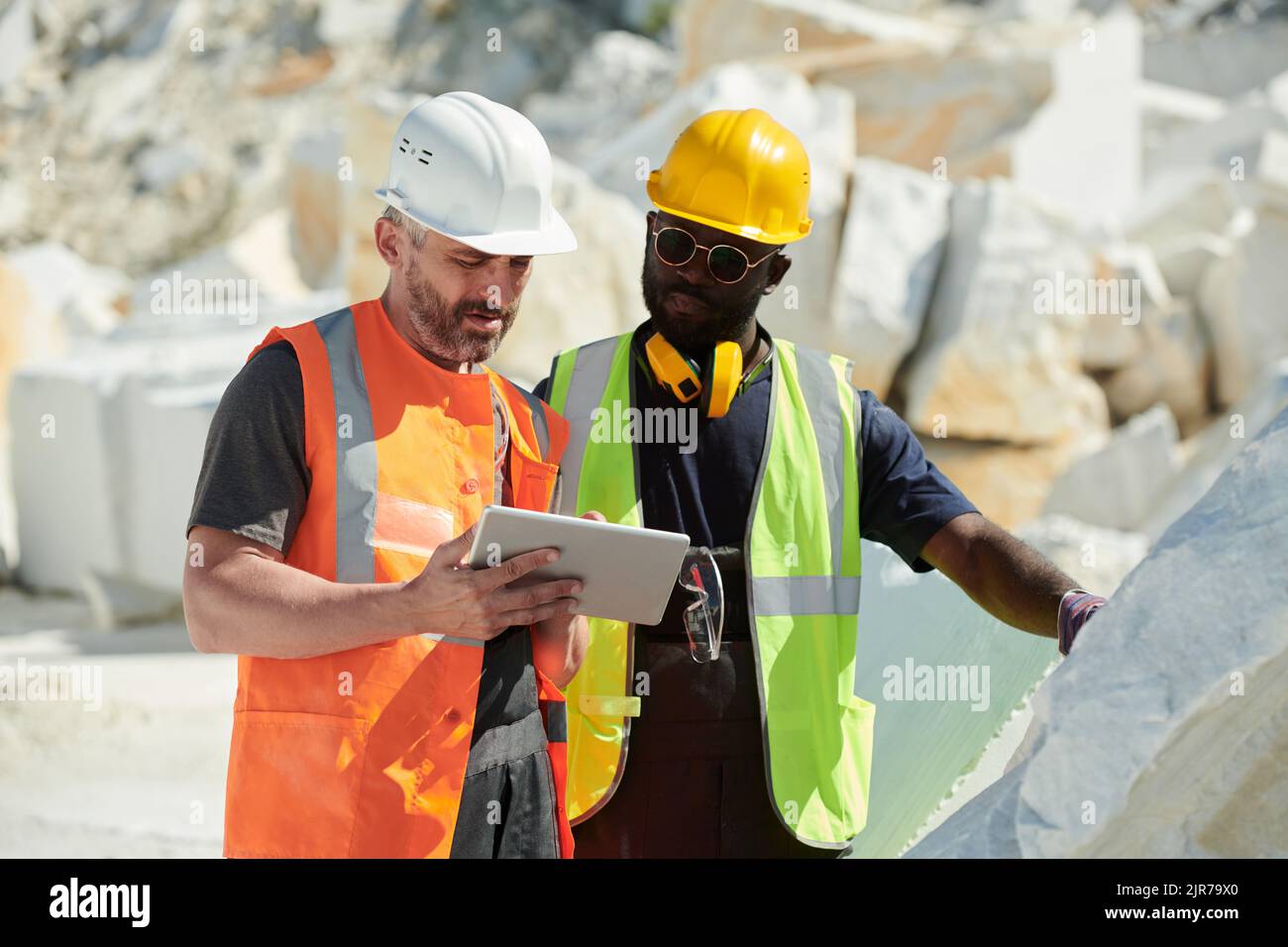 Two interracial male engineers in hardhats and uniform looking at tablet screen while discussing online data or reading manual guide Stock Photo