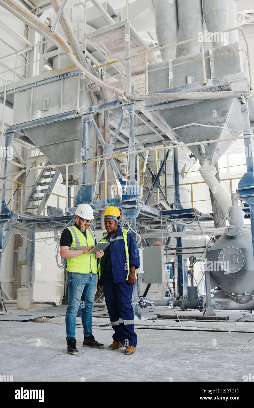 Two young intercultural engineers consulting manual guide in digital tablet while standing in the middle of spacious industrial plant Stock Photo