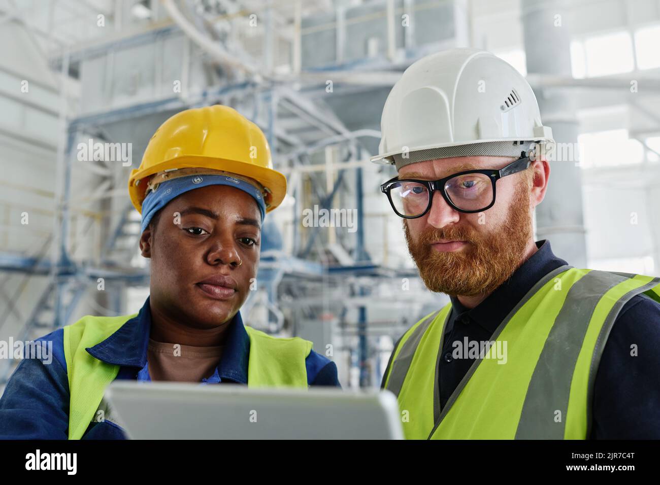 Two workers of modern factory looking through online information at tablet screen while consulting manual guide in workshop Stock Photo