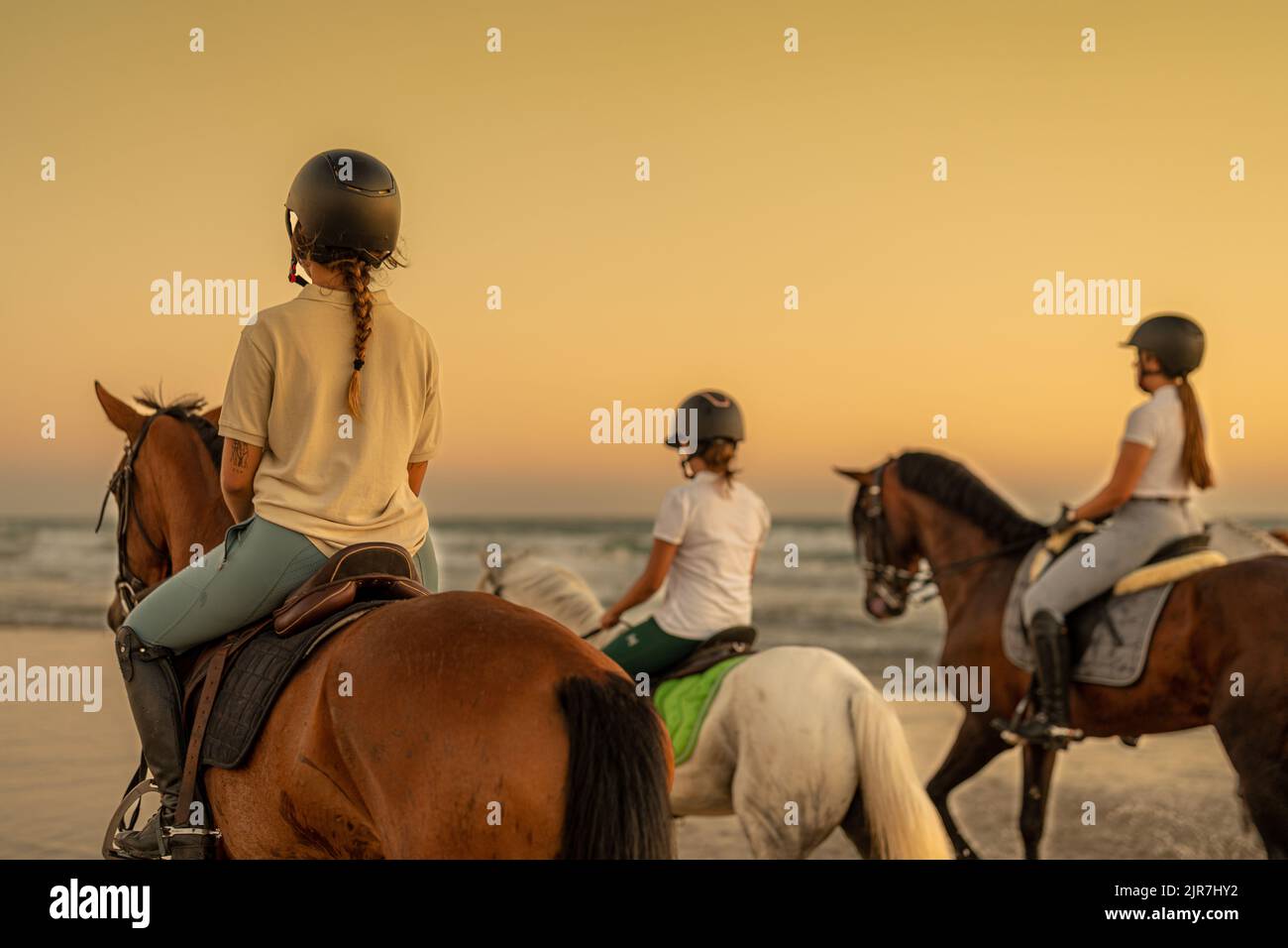 young woman with braid riding a horse on the beach next to 2 young riders Stock Photo