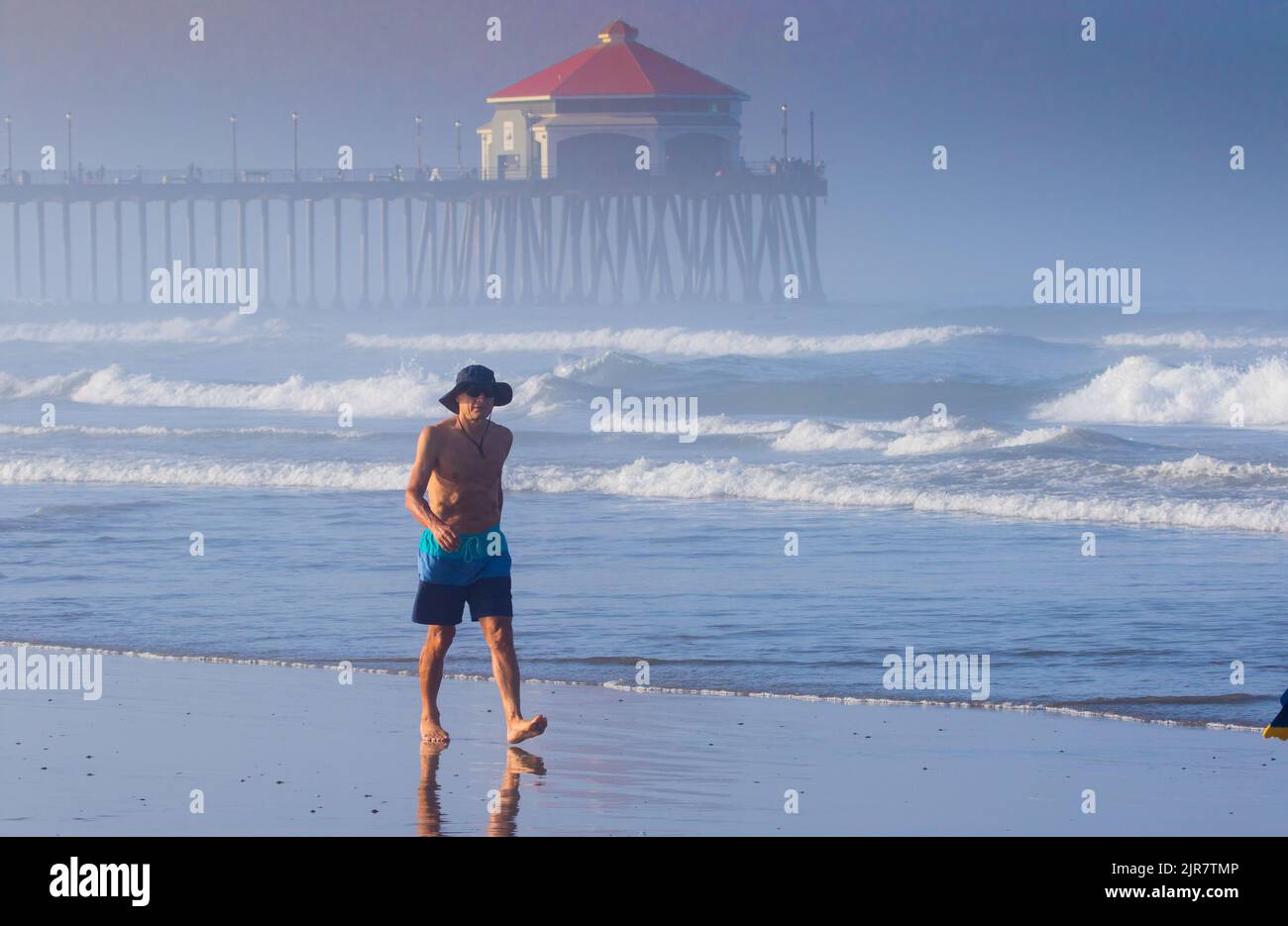 Man walking along the sand at Huntington Beach California wearing a hat and pair of shorts early in the morning with the pier in the background mist . Stock Photo