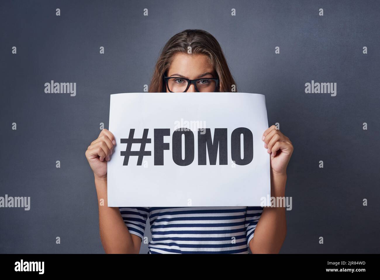 If its happening I want to be part of it. Studio shot of an attractive young woman holding a sign with FOMO printed on it against a gray background. Stock Photo