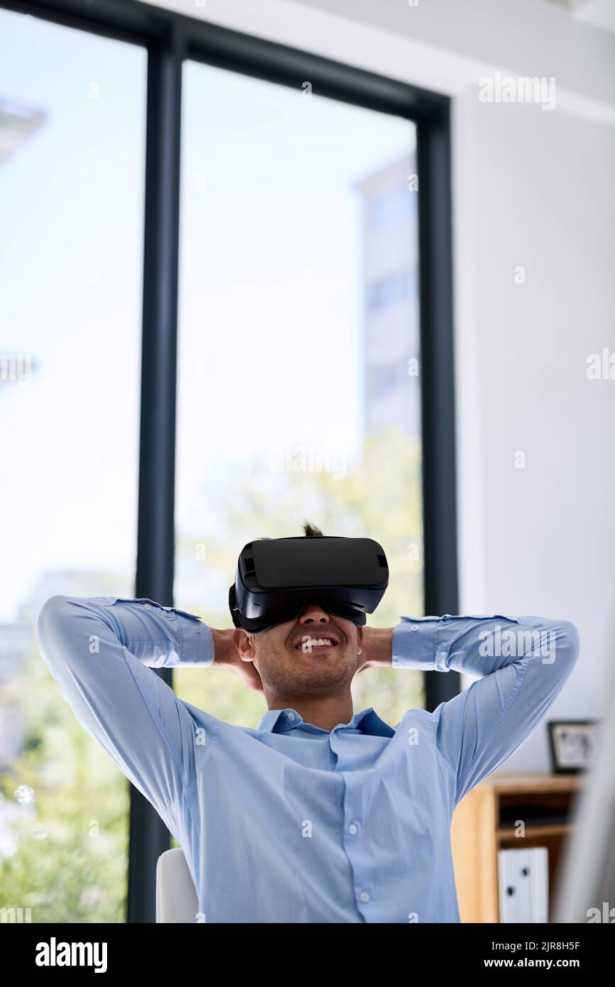 Now he can brainstorm with a beach view. a happy young businessman wearing a virtual reality headset while working at his desk in the office. Stock Photo