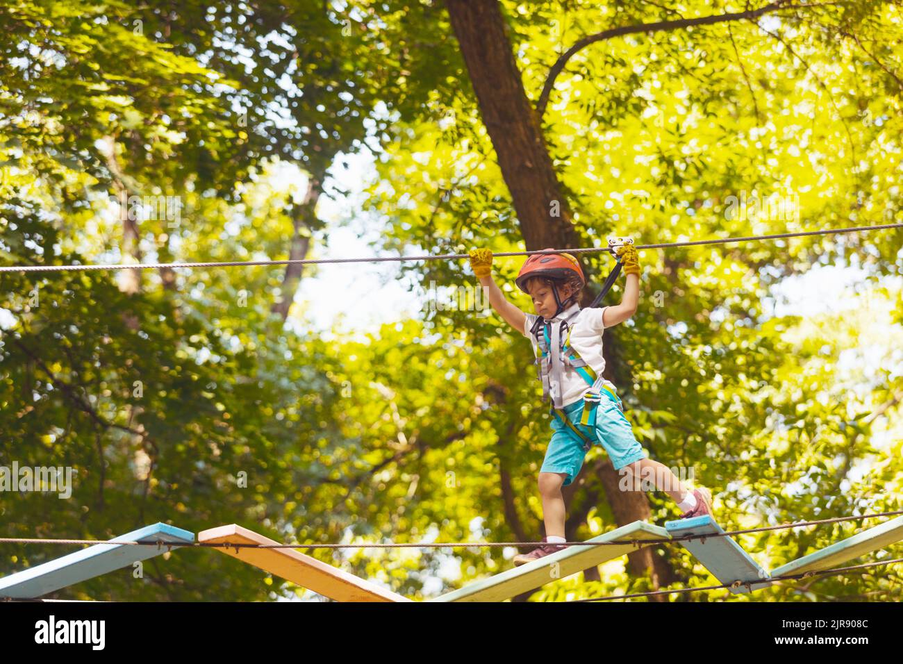 Portrait of a concentrated boy who is climbing in the rope park in summer. He is wearing a protective helmet and gloves Stock Photo