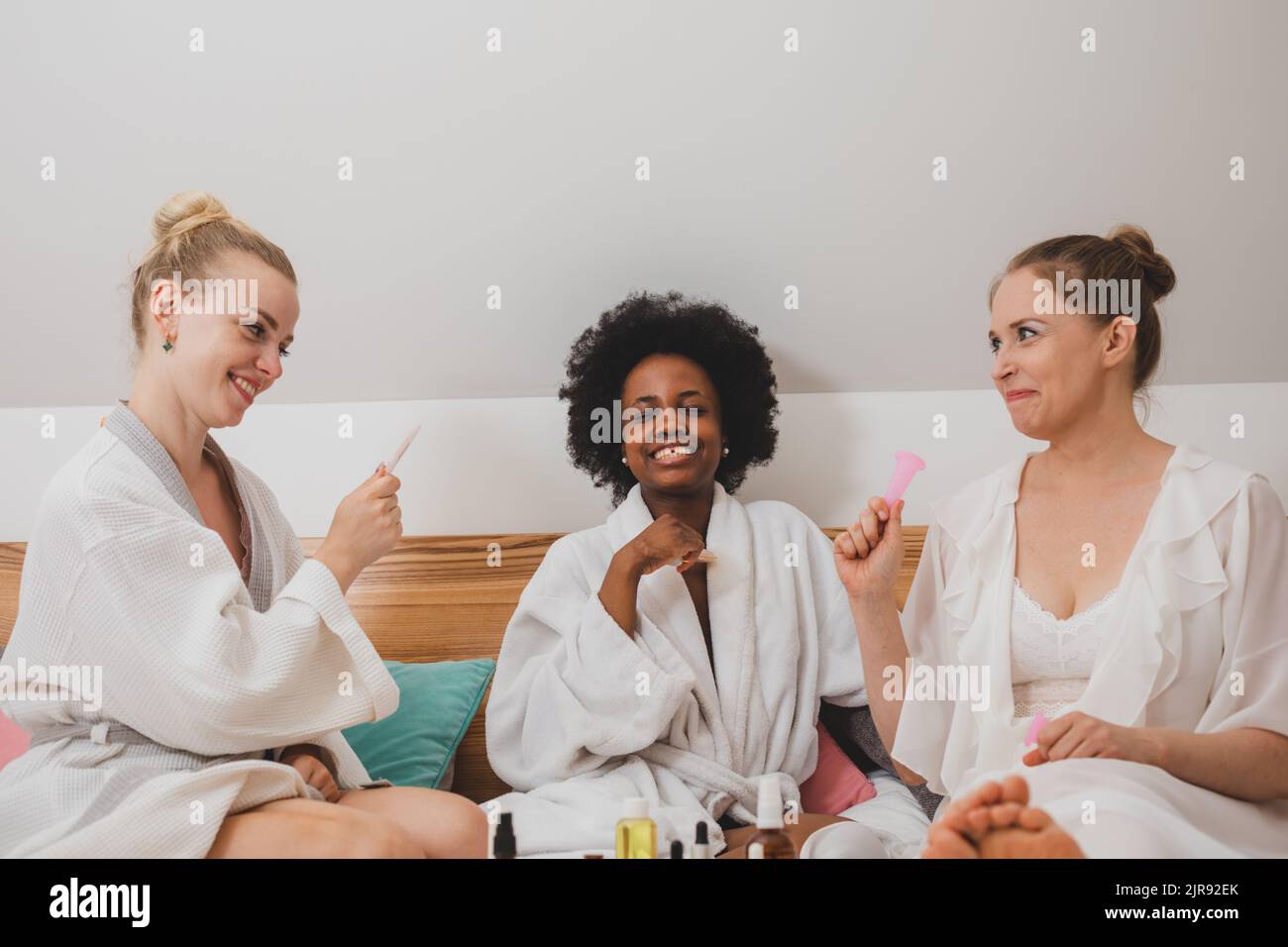 The young women in bathrobes are sitting on the bed and using skincare products Stock Photo