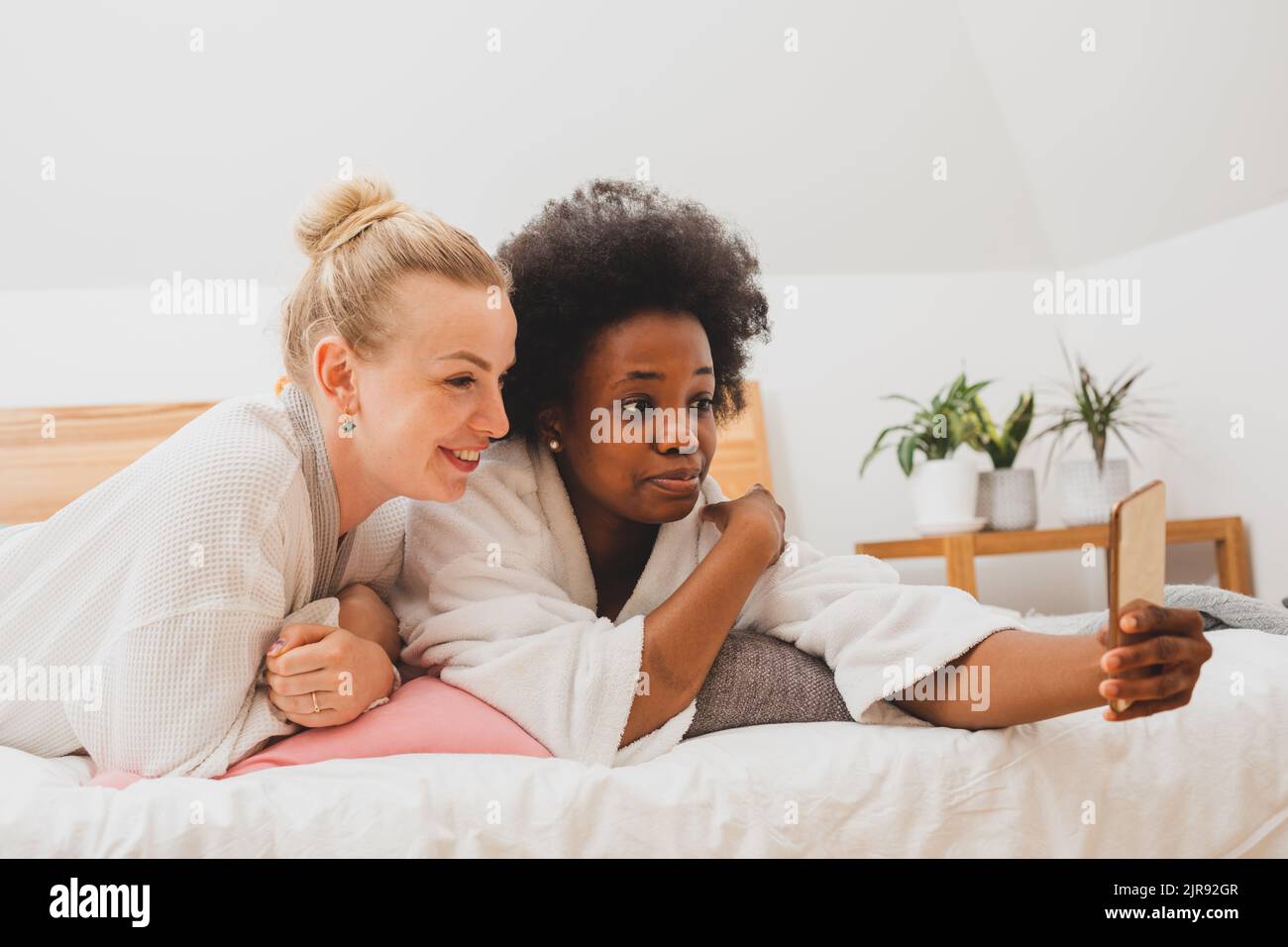 Two young women are lying on the bed and take selfies. The women in bathrobes are resting after spa treatments Stock Photo