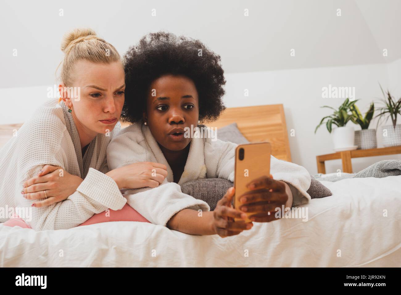 Two young women of different nationalities have a video chat at the spa. The women in bathrobes are lying on the bed and communicating to using a smar Stock Photo