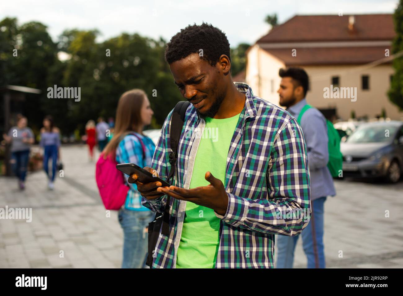 The young african student with a bag outdoors is holding a phone. The man in a shirt is looking for road to home with help navigation Stock Photo