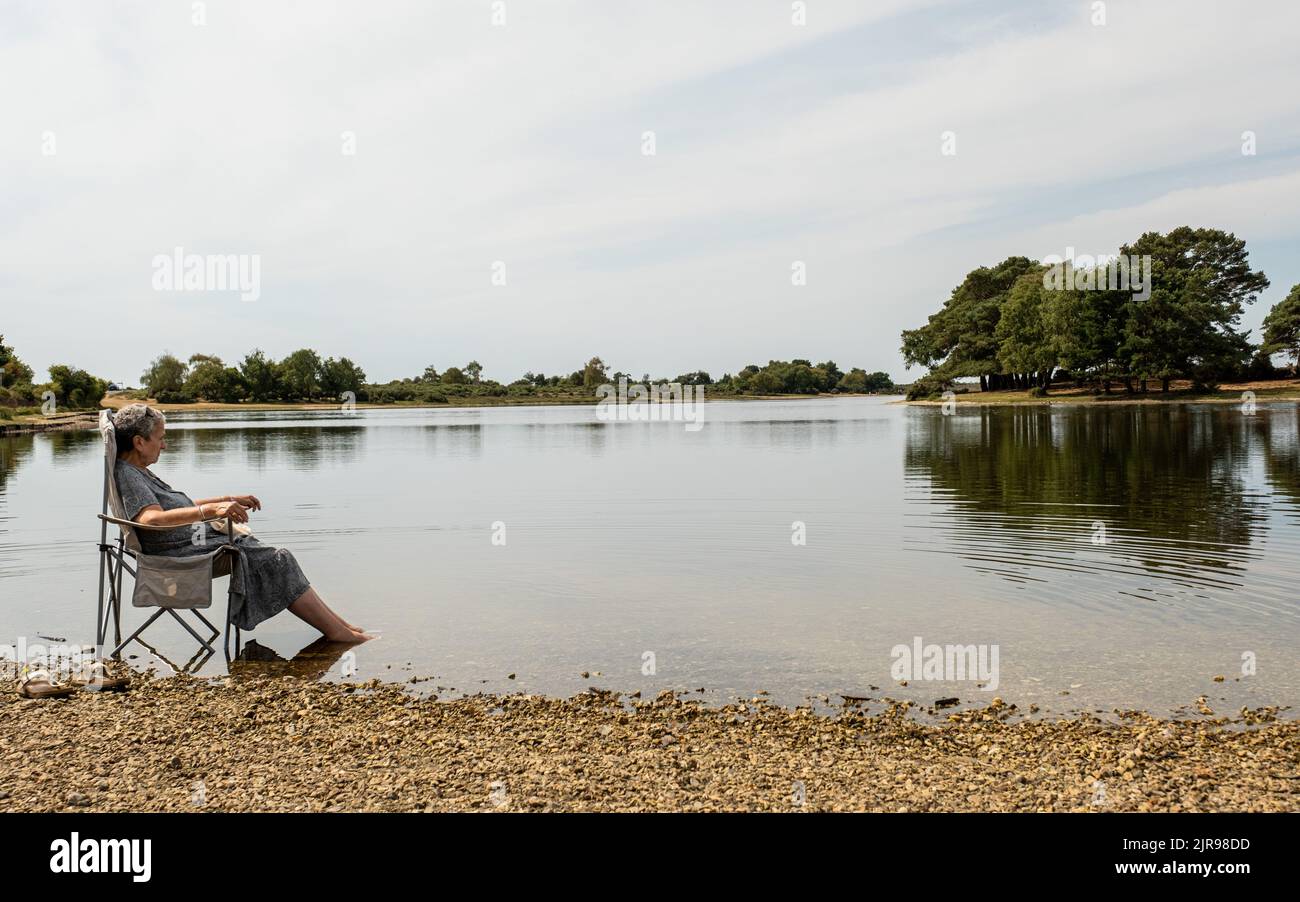 A lady on holiday in New Forest enjoying paddling in a lake whilst sitting on a folding chair in the lake Stock Photo