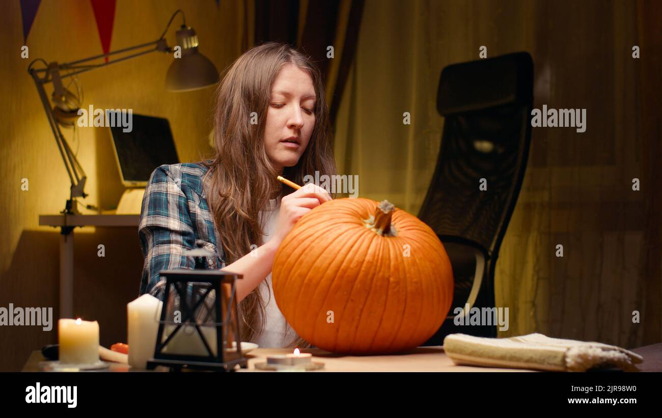 Preparing pumpkin for Halloween. Woman sitting and marking pumpkin with pencil before carving halloween Jack O Lantern at home for her family. Stock Photo