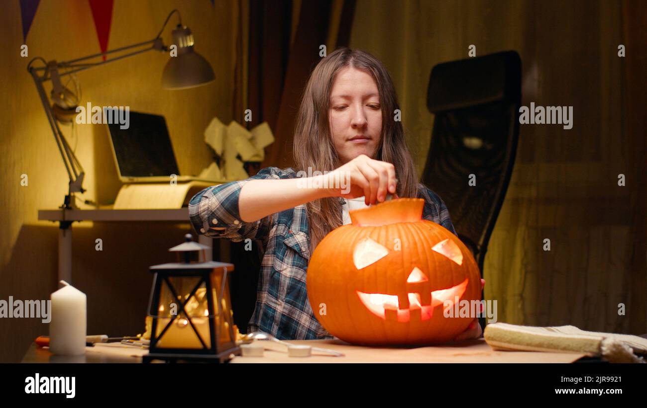 Cutting into pumpkin and taking lid with seeds out. Halloween preparations. Woman sitting and carving with knife halloween Jack O Lantern pumpkin at h Stock Photo